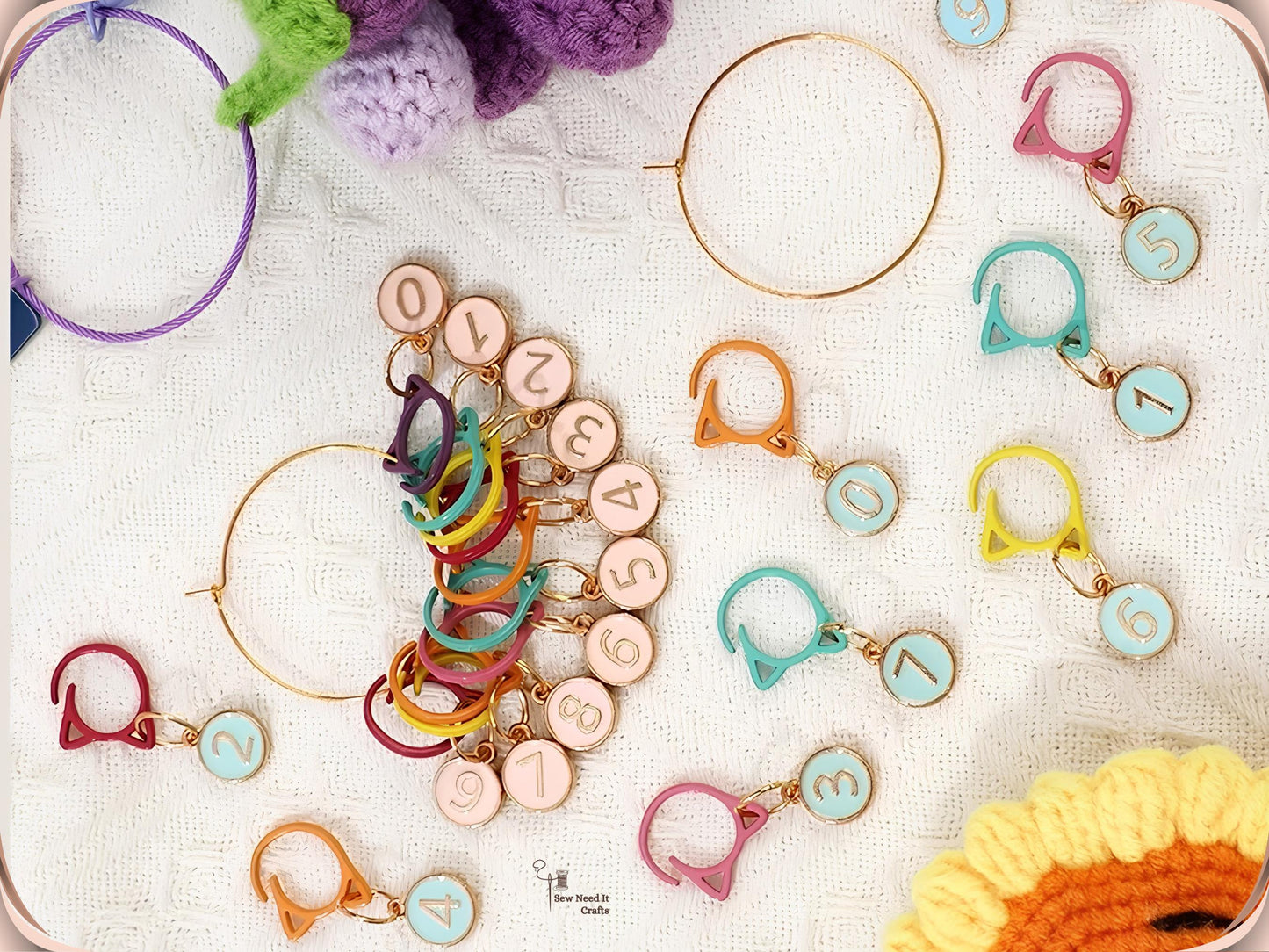 Mixed-colour numbered stitch markers gathered on a gold-tone storage ring on white fabric, with a purple hoop, crochet flowers and yarn in the background