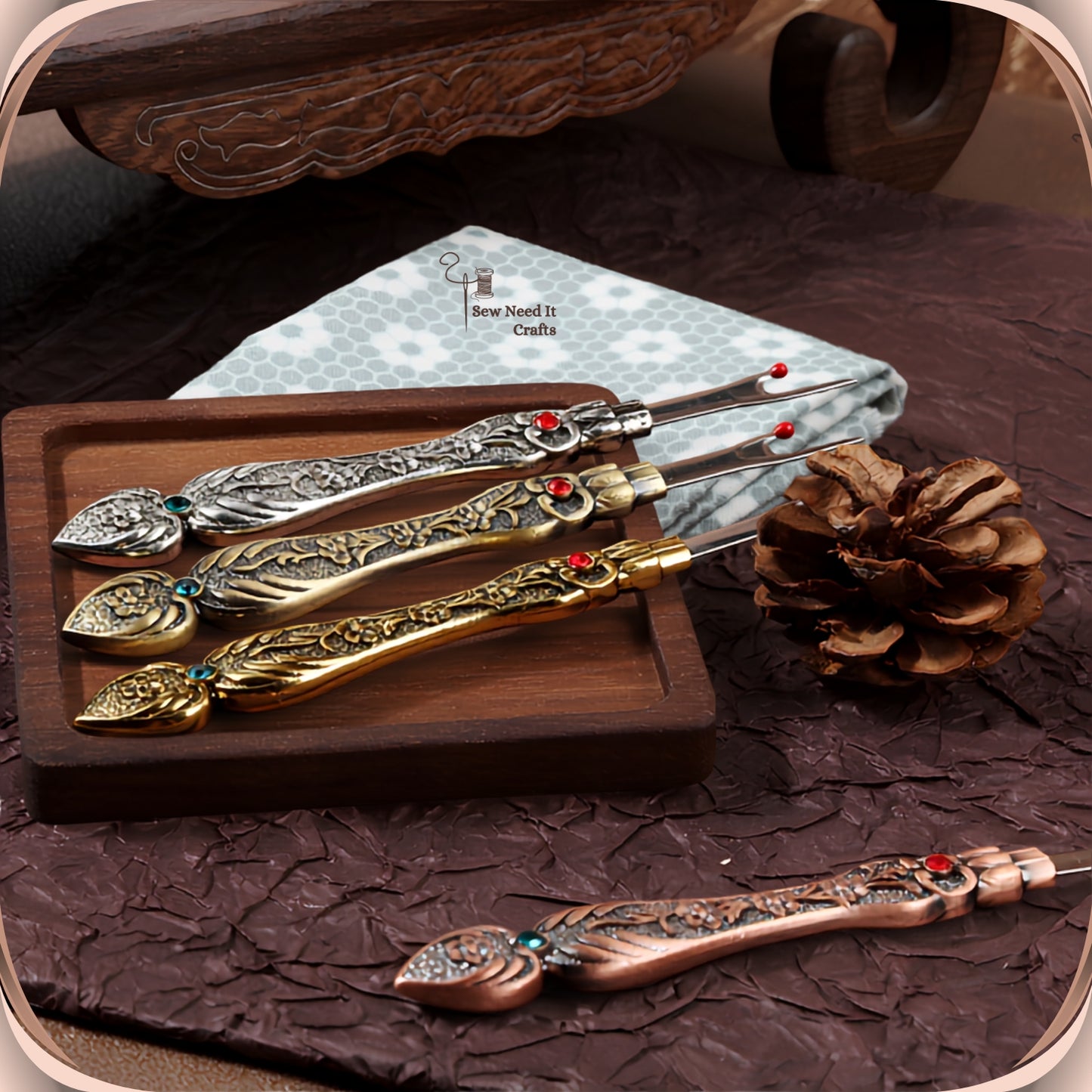 Four Antique Flat seam rippers in silver, bronze and gold on a wooden tray with folded fabric, and a copper one on the surface below