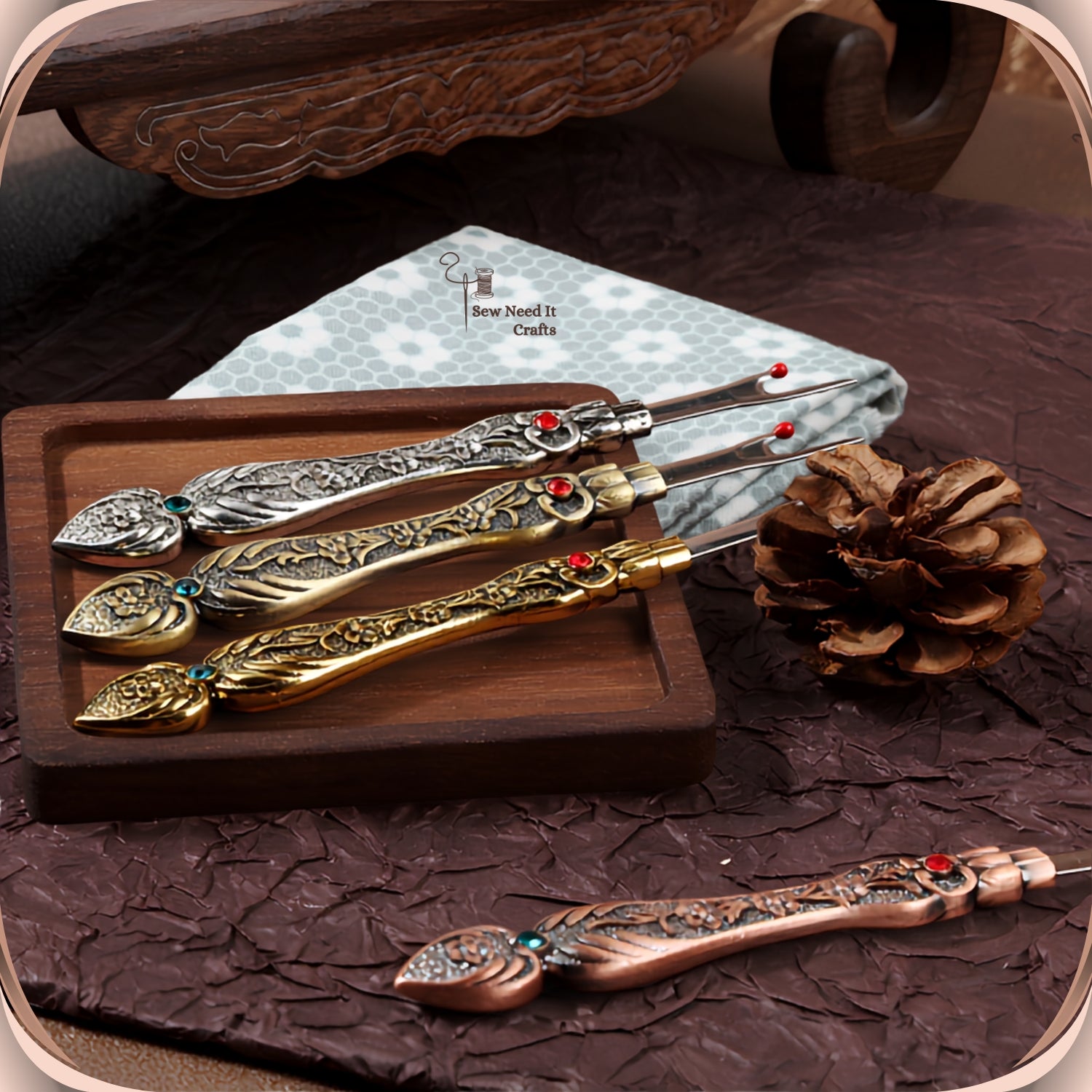 Four Antique Flat seam rippers in silver, bronze and gold on a wooden tray with folded fabric, and a copper one on the surface below