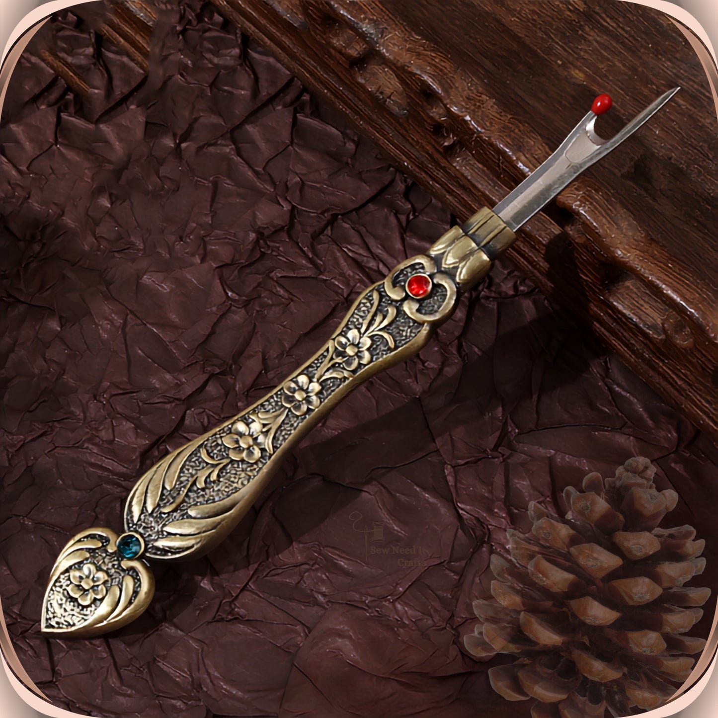 Antique Bronze flat handle seam ripper on a dark plum background, showing the raised blossom engraving, the red crystal at the collar and the teal-green crystal in the tail