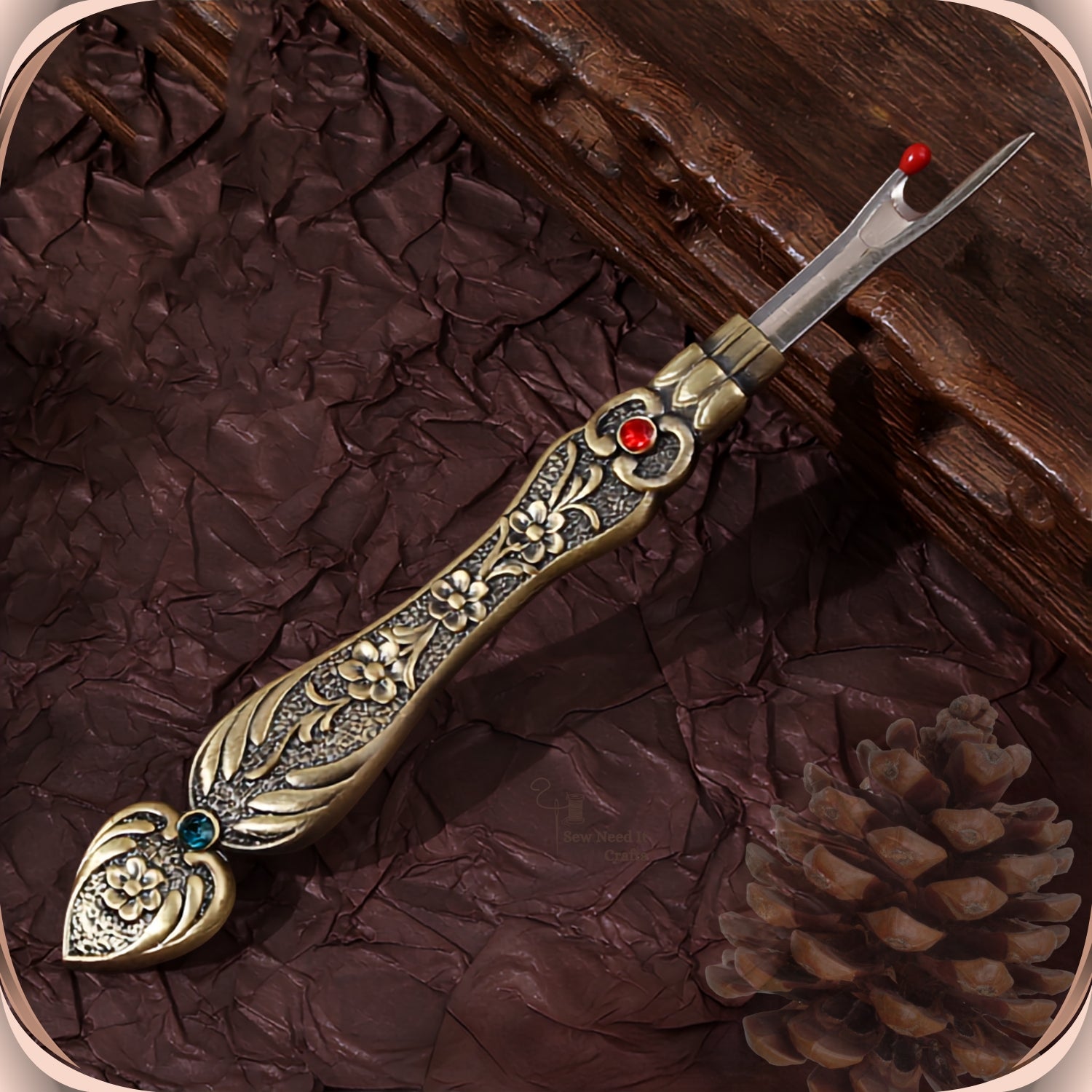 Antique Bronze flat handle seam ripper on a dark plum background, showing the raised blossom engraving, the red crystal at the collar and the teal-green crystal in the tail