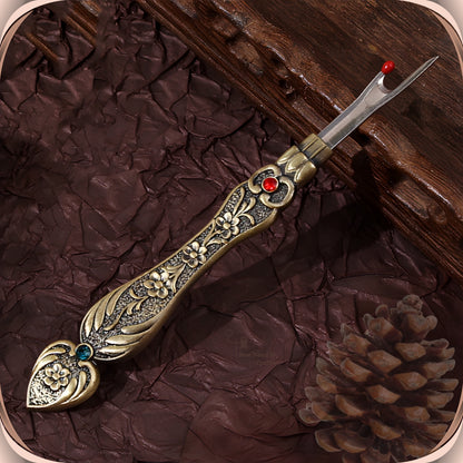 Antique Bronze flat handle seam ripper on a dark plum background, showing the raised blossom engraving, the red crystal at the collar and the teal-green crystal in the tail