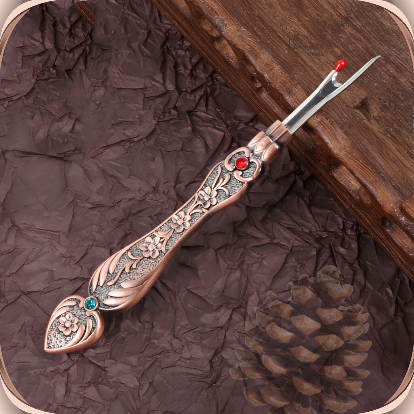 Antique Copper flat handle seam ripper on a dark plum background, showing the raised blossom engraving, the red crystal at the collar and the teal-green crystal in the tail