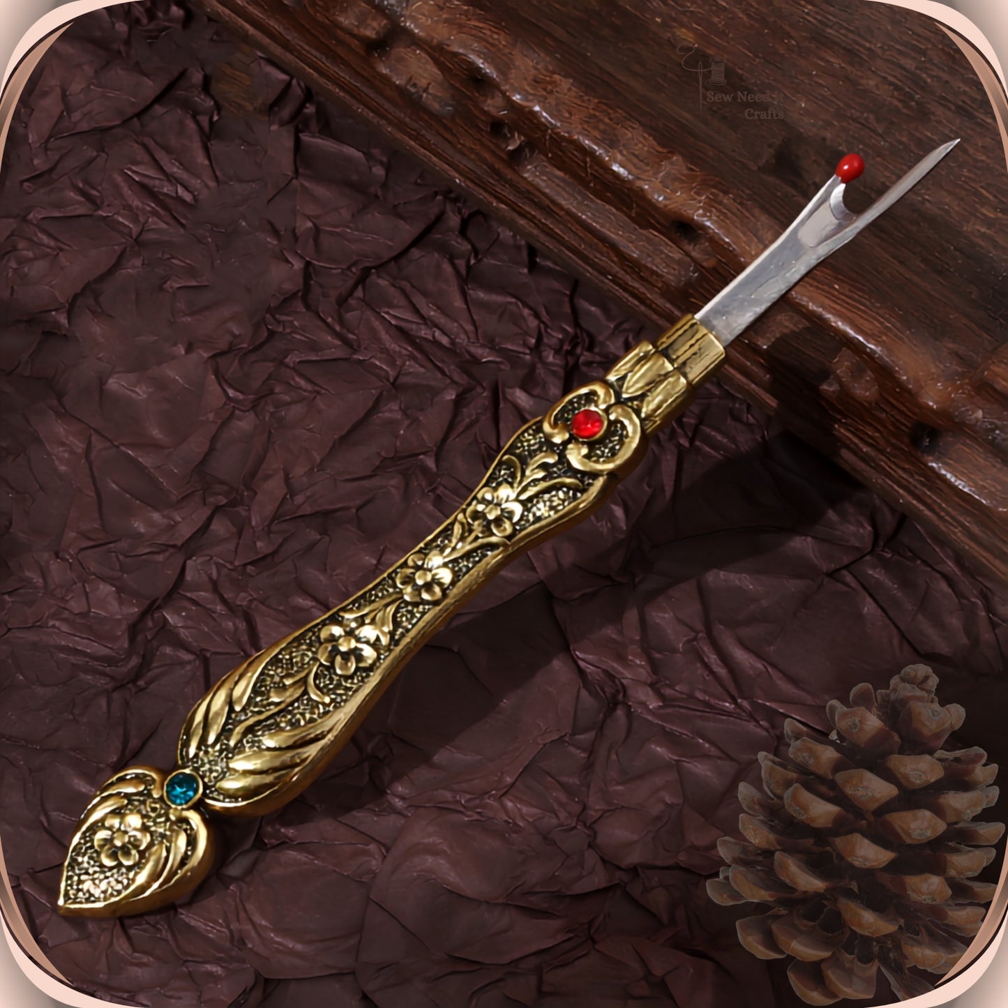 Antique Gold flat handle seam ripper on a dark plum background, showing the raised blossom engraving, the red crystal at the collar and the teal-green crystal in the tail