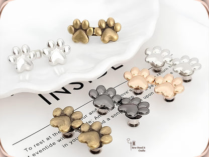 Golden and silver paw waist adjusters fastened on a light denim waistband, with a bronze pair alongside showing the pin backs
