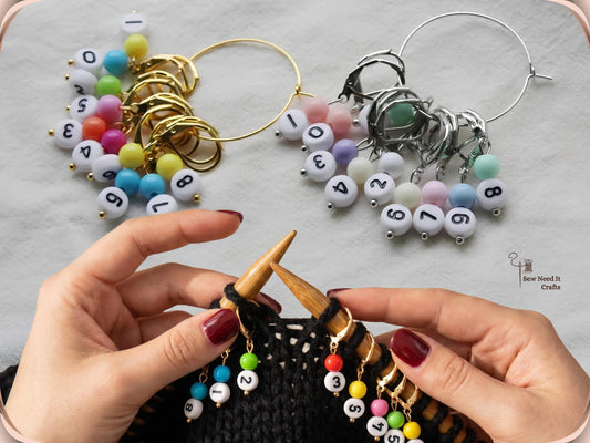 Hands knitting black yarn on bamboo needles with gold numbered stitch markers attached, and the gold and silver sets on their storage hoops above