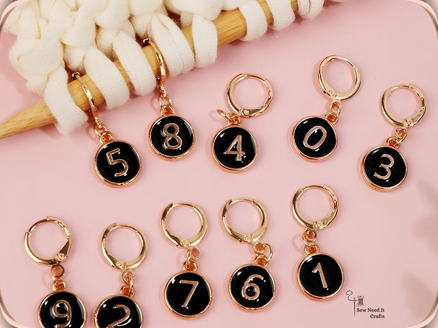 Black enamel numbered stitch markers 0 to 9 with gold clasps arranged on a pink surface beside cream yarn on a bamboo needle