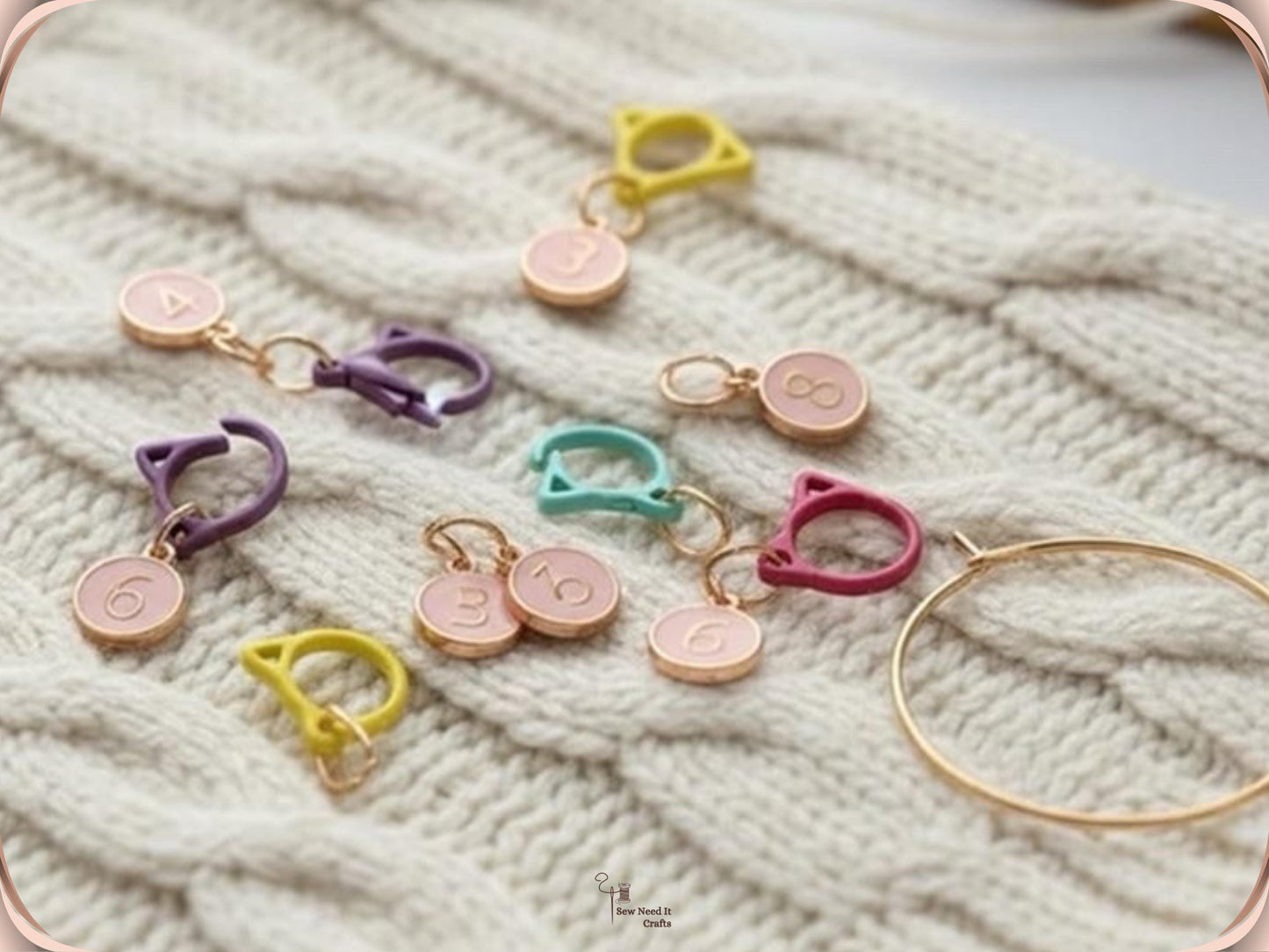 Numbered stitch markers with pink enamel charms clipped into a cream cable-knit fabric, with the gold-tone storage ring resting alongside