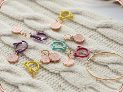 Numbered stitch markers with pink enamel charms clipped into a cream cable-knit fabric, with the gold-tone storage ring resting alongside