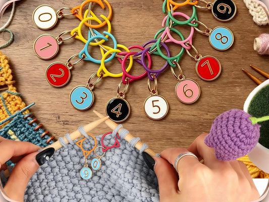 Numbered cat-ear stitch markers clipped onto a grey knitted swatch on the needle, with the full mixed-colour set of ten laid out on a wooden table beside yarn and a crochet flower