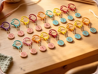 Pink and blue numbered stitch marker sets arranged in rows on a wooden table with knitting needles, yarn and a green knitted swatch