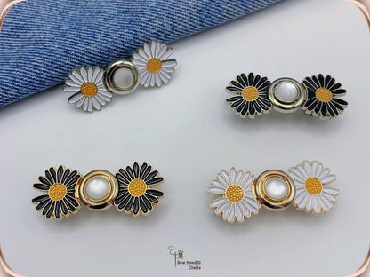 Four assembled daisy waist adjusters in black and white enamel with gold and silver finishes, arranged on white beside folded denim