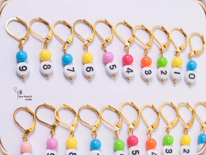 The ten gold-tone numbered stitch markers laid out in order from 9 down to 0, each with a white number bead and a coloured bead
