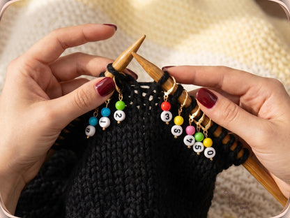 Hands knitting black chunky yarn with gold numbered stitch markers clipped along the bamboo needle