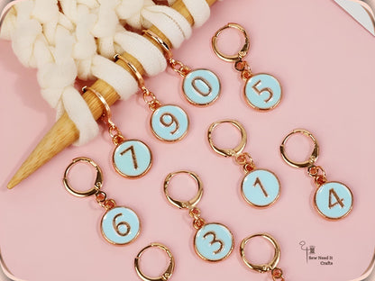 Light blue enamel numbered stitch markers 0 to 9 scattered on a pink surface beside cream yarn on a bamboo needle