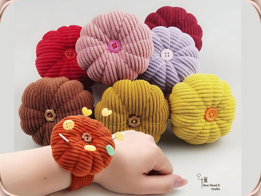 Seven corduroy pumpkin pin cushions in red, dusty pink, dark red, purple, coffee, dark yellow and yellow, with an orange one worn on a wrist holding pins and buttons