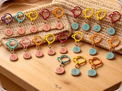 Pink and blue numbered stitch marker sets laid out in rows on a wooden board beside a cream knitted cowl, each set numbered 0 to 9 on assorted-colour cat-ear clips