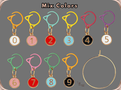 The Mix Colors set of ten numbered stitch markers, 0 to 9, with charms in white, pink, red, blue and black on assorted coloured cat-ear clips, shown next to the gold-tone storage ring
