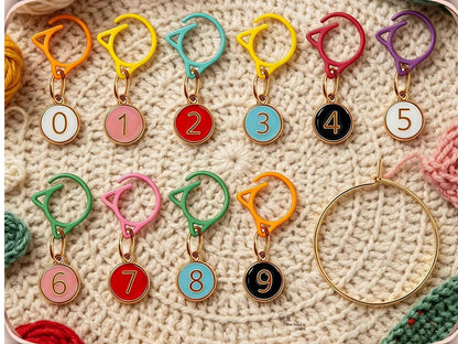 Mixed-colour numbered stitch markers 0 to 9 arranged on a cream crochet blanket with the gold-tone storage ring alongside