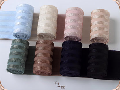 Eight rolls of 2 inch wide striped elastic band partly unrolled, shown from above in light blue, cream, pink, apricot, sage, mauve, navy and black