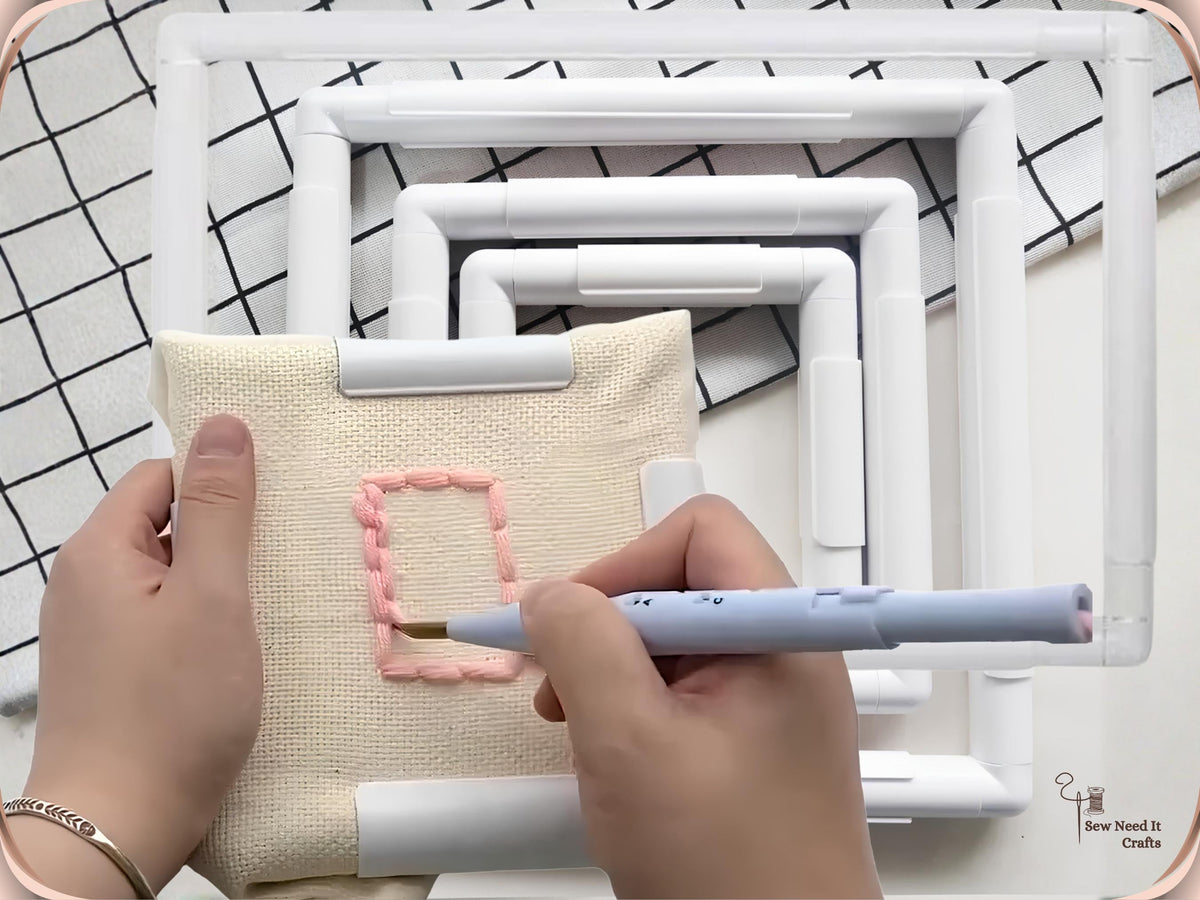 Blue punch needle working pink yarn into cream fabric held in a white square snap frame, with larger frames behind