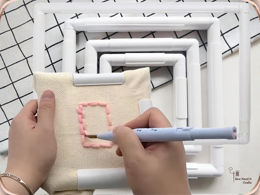 Blue punch needle working pink yarn into cream fabric held in a white square snap frame, with larger frames behind