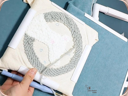 Punch needle project on cream cloth clamped in a large white snap frame, blue punch needle in hand