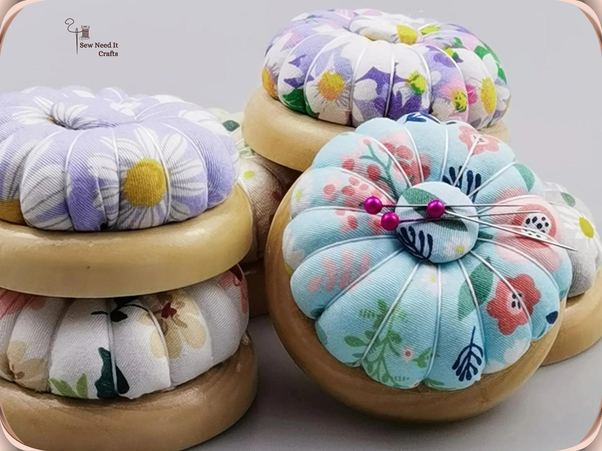 Light blue floral pin cushion with pins in its magnetic center, beside stacked purple and white daisy pin cushions on wooden bases