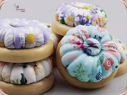 Light blue floral pin cushion with pins in its magnetic center, beside stacked purple and white daisy pin cushions on wooden bases