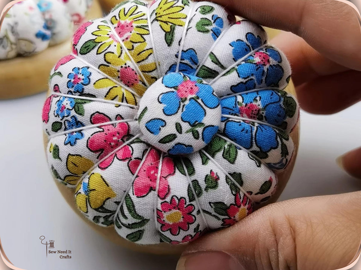 Hand holding a white pin cushion printed with blue, pink and yellow flowers, showing its pumpkin sections