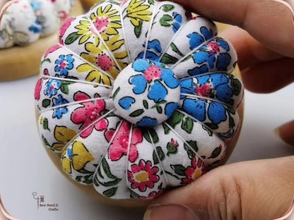 Hand holding a white pin cushion printed with blue, pink and yellow flowers, showing its pumpkin sections