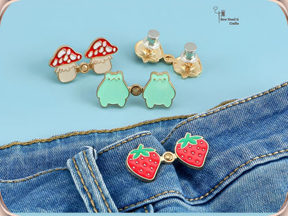 Mushroom and frog waist pins with silver back caps above a strawberry pair snapped onto a jeans waistband