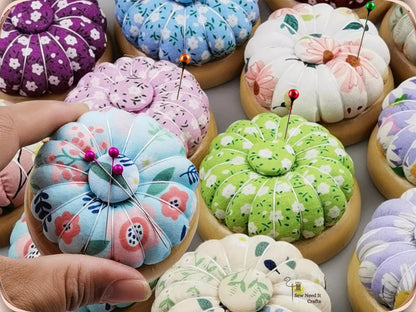 Hand holding a light blue floral pin cushion with pins and needles, among other wooden-base pumpkin pin cushions