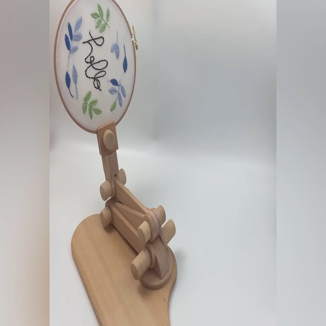 Video demonstrating the beechwood embroidery hoop stand: the hoop being unclamped and re-clamped by hand, the arm folding and extending through its height range, 360 degree rotation at the clamp and base, and the stand used as a sit-on lap stand on a chair