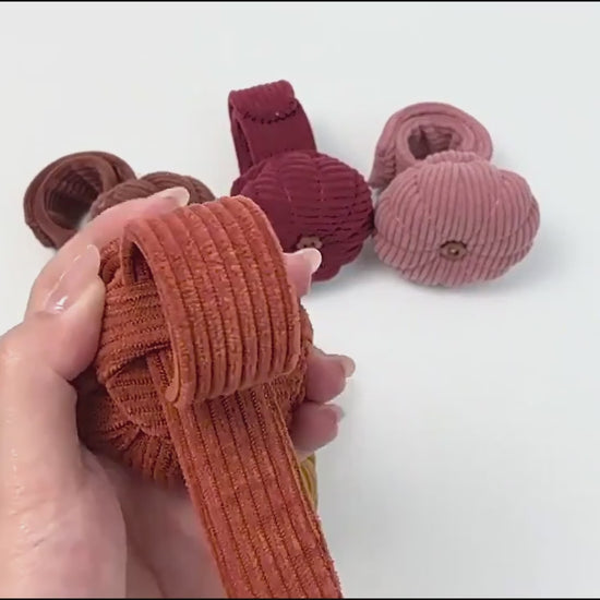 Hands demonstrating the pumpkin wrist pin cushion: the corduroy band is shown lying flat, then wrapped and snapped closed around a wrist, and finally holding several button-head sewing pins while worn