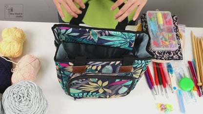 Video of the crochet and knitting project bag: yarn threaded out through one of the four holes in the top panel, several balls loaded inside, the top zipped shut, the front flap dropped to show a row of crochet hooks under an elastic band, and straight knitting needles standing in the outside side pockets