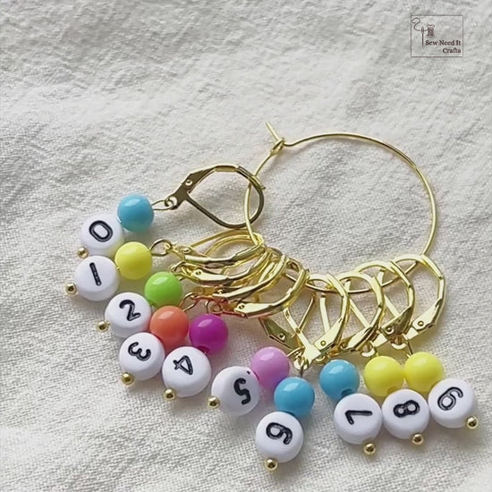 Video showing the gold set of numbered stitch markers on its storage hoop, a leverback marker opened and snapped shut by hand, and a marker clipped onto a bamboo knitting needle mid-project