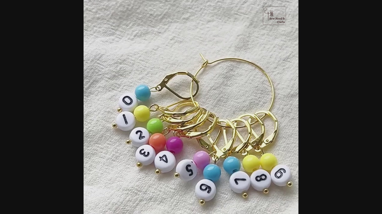 Video showing the gold set of numbered stitch markers on its storage hoop, a leverback marker opened and snapped shut by hand, and a marker clipped onto a bamboo knitting needle mid-project