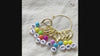 Video showing the gold set of numbered stitch markers on its storage hoop, a leverback marker opened and snapped shut by hand, and a marker clipped onto a bamboo knitting needle mid-project